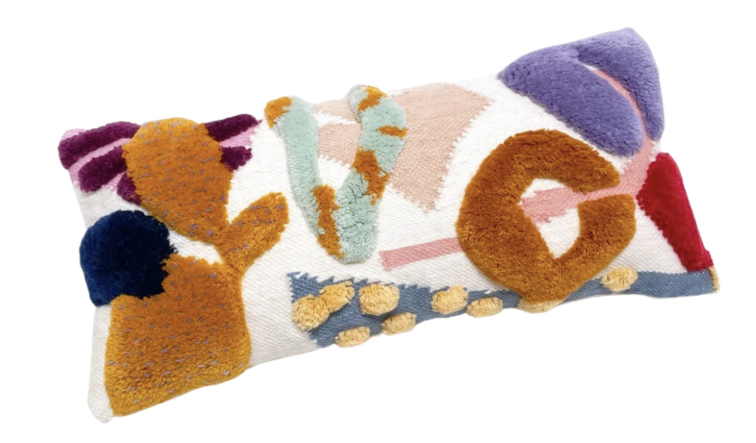 A high-angle shot of a rectangular lumbar pillow with a multi-textured, abstract design on an off-white background. The design elements are colorful and appear to be tufted or embroidered with raised surfaces. From left to right: On the far left, a purple, irregular shape with a fluffy texture. Adjacent to it, a dark blue, rounded, amorphous shape. Next to the blue shape, a large, prominent, bright orange shape that resembles a cactus or an abstract hand with two fingers pointing rightwards. This shape is very textured and raised. To the right of the orange shape, the letters "V" and "G" are visible, also highly textured and raised. The "V" is light teal or mint green with some darker flecks. The "G" is a rich, warm brown or rust color, forming a large, thick letter. Surrounding and between these main elements are various smaller, softer-colored abstract shapes: a light pink, possibly semicircular shape above the "V," and thin horizontal lines of light pink below the "V" and "G." In the bottom center, below the "V" and "G," is a patch of muted blue with several raised, irregularly shaped yellow or gold dots or pebbles. On the far right, partially visible, is a section of red fabric with a slightly textured appearance, and above it, a large, irregular lavender or light purple shape with a fluffy texture. The pillow has a playful, artistic, and tactile aesthetic, suggesting a mix of abstract forms and perhaps some hidden letters.